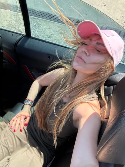A young woman with blonde hair, wearing a pink baseball cap and a brown tank top, is seated in the passenger seat of a car. Her hair is blowing across her face due to wind. She is wearing a watch and several bracelets on her left wrist, and two rings on her right hand, with bright red nail polish. The interior of the car is visible, including black leather seats with red accents and a dark dashboard. Outside the car window, a paved area with drainage grates is visible. The scene appears to be outdoors on a sunny day. The location is the city of Dzegvi, Georgia.
