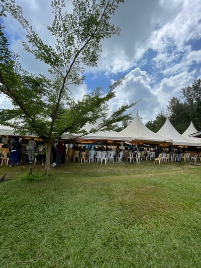 This image, taken in Makuyu, Kenya, depicts an outdoor gathering under a partly cloudy sky. A large tree with green foliage dominates the left side of the foreground, its branches extending towards the center. To the right of the tree, a series of white canopy tents are erected on a vast green grassy field.
Underneath these tents, numerous individuals are seated on rows of plastic chairs, which are a mix of white and beige/brown. The arrangement of chairs suggests an organized event such as a meeting, seminar, or social function. People appear to be attentive, though specific expressions are not discernible from this distance.
The sky overhead is bright blue with scattered, large white cumulus clouds, indicating it is daytime with fair weather conditions. Additional trees are visible in the background, suggesting a natural or open environment surrounding the event area. The overall scene conveys an atmosphere of an organized outdoor event taking place in a natural setting. No visible text or branding can be identified.