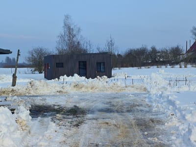 The image depicts a winter scene in Skudras, Latvia. In the foreground, a partially cleared dirt or gravel path extends from the bottom of the frame towards the midground. The path is wet, slushy, and muddy in areas, exhibiting visible tire tracks, and is flanked by irregular piles of dirty snow. In the midground, a dark-colored, single-story rectangular building with vertical dark siding is situated. The building features two dark-framed windows and a dark-framed door on its visible side, with a prominent vertical red accent stripe on its left facade. Piles of snow surround the base of the structure. To the immediate left of the path, a weathered wooden post stands. The background consists of a vast, snow-covered field dotted with numerous bare deciduous trees and shrubs. A faint fence line is visible between the foreground snow and the field. The sky above is clear and pale blue. No individuals are present in the scene.
