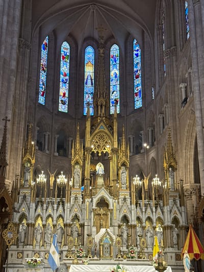 The interior of a grand cathedral showcases intricate architectural details. Stained glass windows in varying shades of blue depict religious scenes. Elaborate altars, adorned with statues, gold accents, and lights, line the wall. The vaulted ceilings and stone columns contribute to the cathedral's impressive scale and ornate design. Flags and other decorations add color to the sacred space.