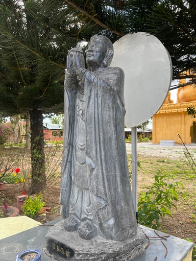 An outdoor scene in Đăk Mar, Vietnam, depicts a dark grey stone statue of a robed figure positioned centrally. The figure stands with hands clasped together at chest level, looking slightly upwards, and wears a cap-like head covering. The statue rests on a dark grey stone base, which bears the inscription "TO BA-TU-MAT" on its front edge. A small, partially visible blue bowl containing dark material sits on the base to the left of the inscription.
Behind the statue, a large pine tree with green needles extends into the upper left quadrant of the frame. To the left of the statue, a garden area features red and yellow flowers, various green plants, and potted plants, with smaller red-roofed buildings visible in the distance. To the right of the statue, a large, round, white panel or disk stands upright, partially obscuring a prominent golden temple building with multi-tiered, curved roof details. The ground around the statue is a mix of sparse green grass and bare earth, with fallen pine needles or small branches scattered on the statue's base and the ground. The scene is illuminated by natural daylight. No individuals are present in the image.