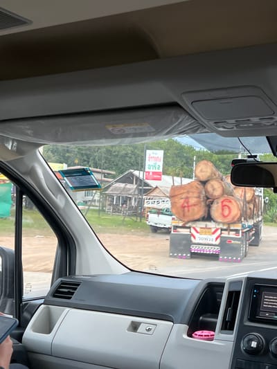 The image shows a view from inside a vehicle, looking out at a road scene in Ban Ko Pru, Thailand. A large truck carrying a load of logs is the primary subject in the outdoor scene. The logs are substantial in size, and some have red markings on them, including what appears to be a "4" and a "6". The truck has reflective safety markings on its rear.

The scene is set during the day, with daylight visible. The weather appears to be overcast or slightly hazy, as the light is diffused. In the background, there are some buildings with thatched or corrugated roofs, suggesting a rural or semi-rural setting. There are also trees and greenery visible, indicating an outdoor environment.

Inside the vehicle, the dashboard and parts of the interior are visible, including air vents and a screen. A pink air freshener is placed in one of the compartments. There is also a sticker on the windshield with some text, and another sticker that appears to be a warning label. The overall impression is a snapshot of travel, with the vehicle's interior framing the external view.