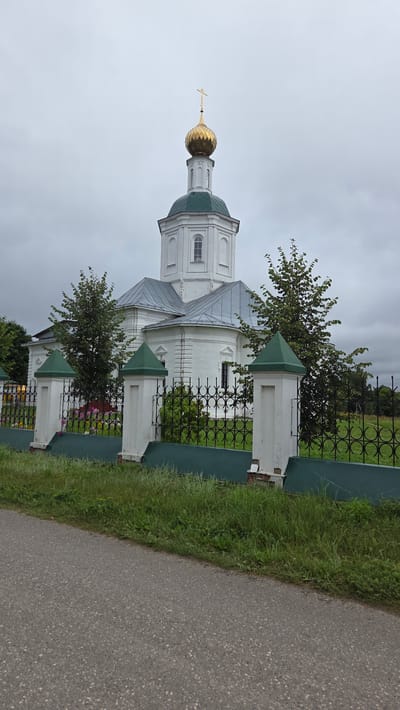 The image shows a white orthodox church with a green roof and a golden dome topped with a cross. The church is surrounded by a black metal fence with white pillars topped with green pyramid-shaped caps. A paved road is visible in the foreground, with grass and some flowers behind the fence. Trees are scattered around the church. The sky is overcast. The church is located in the city of Novo-Talitsy, Russia.
