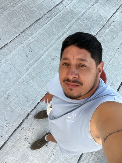 An adult male subject is depicted in an overhead, slightly angled photograph, looking directly at the camera. He has dark, short hair, a mustache, and a short beard. A small silver-colored hoop earring is present in his left ear. He is wearing a light blue sleeveless t-shirt with a partially visible white circular graphic containing text, including "COMPANY" and "PROVISIONS." White shorts and dark olive green sneakers are visible on his lower body. A script tattoo spelling "Adriana" is visible on his left bicep. A small segment of a red object, possibly a chair, is visible behind his head. The ground surface beneath him is light-colored, textured pavement, likely composed of rectangular tiles or stamped concrete in a patterned arrangement, with darker linear elements indicating seams or cracks. The scene is brightly lit. The location is Bernal Este, Argentina.