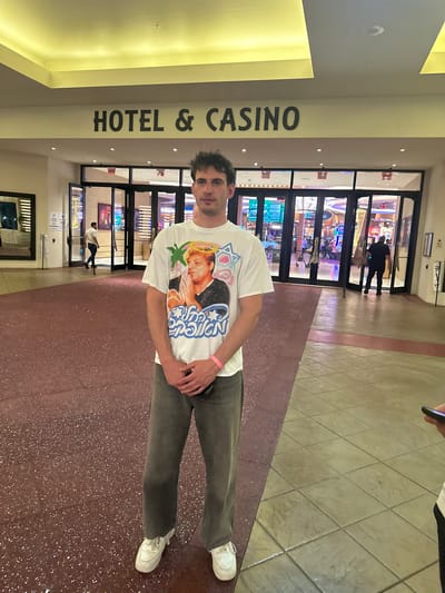 A man stands inside the Seminole Hard Rock Hotel & Casino in Hollywood Seminole Reservation, United States of America. He is wearing a white t-shirt with a graphic print, gray pants, and white sneakers. He has short dark hair. The casino entrance is visible behind him, with glass doors and people walking through. The interior flooring is a speckled reddish-brown. A sign reading "HOTEL & CASINO" is visible on a wall to the left. The lighting is bright and artificial.