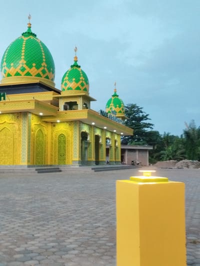 This image depicts the brightly illuminated Noor Taufiq mosque located in Aluekeujruen, Indonesia, under a light blue, partially clouded sky, suggesting dusk or early morning.

In the immediate foreground, to the right, a prominent rectangular yellow pillar with a square, illuminated light fixture on its top stands on a paved surface composed of grey interlocking pavers. These pavers extend across the entire forecourt of the mosque.

The mosque itself is a multi-tiered structure characterized by a striking color palette of bright yellow and green. Two main domes are visible on the left side of the building: a larger dome and a slightly smaller one to its right. Both domes feature a distinctive checkerboard pattern of bright green and yellow segments and are topped with golden finials. A third, smaller dome is partially visible further to the right and deeper into the scene.

The mosque's facade is predominantly bright yellow, adorned with intricate, possibly floral or geometric, patterns in a lighter green hue. Multiple arched windows or decorative recesses are visible on the walls. White light fixtures are embedded along the eaves and within some architectural elements, casting light onto the yellow walls and highlighting the green patterns. Steps lead up to the main entrance area of the mosque.

In the midground, behind the mosque's right side, significant green vegetation, including tall trees and what appear to be palm trees, is visible. To the right of the mosque and partially obscured by trees, a simple, unpainted building, possibly constructed of concrete or brick, is present. Nearby, piles of earth or construction materials are evident. Faint, blurry figures, possibly two or three individuals, are minimally discernible near the distant building.
