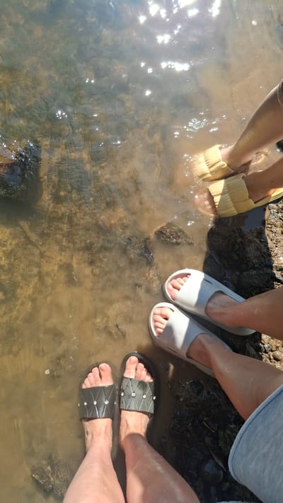 The image captures a close-up, overhead view of four feet submerged in shallow, murky water at the edge of a body of water, likely a river or lake. Two pairs of feet are visible. The pair on the left wears black slides with a diamond-patterned strap adorned with small, clear embellishments. The pair on the right wears light gray slides. Above these, another pair of feet is visible wearing pale yellow slides. The water is unclear, with a sandy or muddy bottom visible beneath the surface. Sunlight reflects brightly off the water's surface, creating shimmering highlights. The bank on the right side of the image is composed of dark, wet soil and rocks. The scene suggests a recreational activity like wading or cooling off in the water on a sunny day. The location is specified as Дзяржынскі сельскі Савет, Belarus.