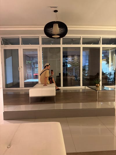 An interior view shows a man seated, casually posed on a white bench, holding a phone. He wears a tan outfit and white cap. Behind him, a multi-paned window reveals an outdoor setting. To the left hangs a unique, spherical black and white pendant lamp. The room features a mix of grey and white surfaces.