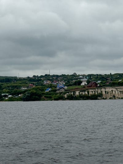 The image depicts a wide shot of a body of water in the foreground, with a small town nestled on a hillside overlooking the water in the background. The sky is overcast and gray. The town appears to be located in Krasnovidovo, Russia.
The town is characterized by a mix of residential buildings, with some appearing to be houses and others larger structures with blue roofs, possibly a church or public building. The buildings are situated among lush green trees that cover the hillside. A prominent feature is a steep, rocky cliff face that separates the town from the water's edge. The water itself is a deep gray, with ripples indicating a gentle current.
There are no people visible in the image. The scene is static and peaceful, suggesting a calm, ordinary day in this Russian town. The overall impression is one of a quiet, rural landscape.