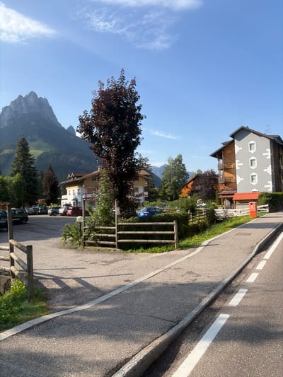 This is a sunny, outdoor, eye-level, medium shot of a street scene in Pera, Italy. The scene features a parking lot with several cars, a wooden fence, a sidewalk, and a road. In the background, there are buildings, trees, and mountains. The sky is blue with some clouds. The overall atmosphere is peaceful and serene.