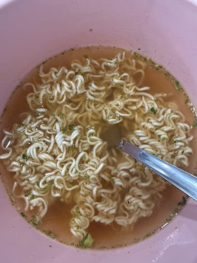The image displays a close-up, top-down view of a light pink or peach-colored bowl containing rehydrated instant ramen noodles in a translucent yellowish-brown broth. Scattered green flakes, likely dried herbs, and smaller reddish specks, possibly chili flakes, are visible within the broth and adhering to the curly, light-colored noodles. A silver-colored metal spoon is partially submerged in the broth, positioned diagonally from the lower right toward the center of the bowl. No people are visible. The scene is located in Madeley, United Kingdom.