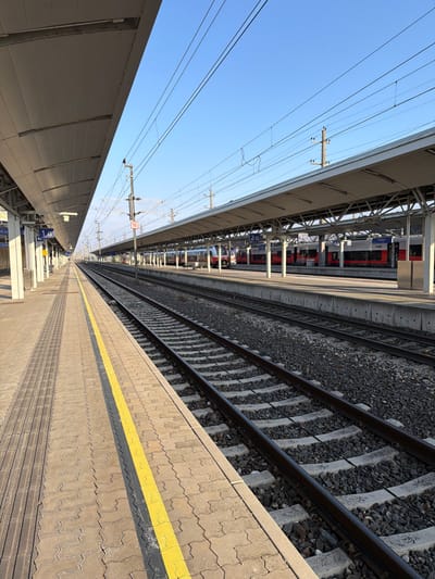 The image displays a deserted train station in Amstetten, Austria, under a clear blue sky. The perspective is from a concrete platform, looking eastward down multiple sets of railway tracks. In the immediate foreground, a yellow safety line demarcates the platform edge, which is surfaced with interlocking pavers and includes tactile paving. Two sets of active railway tracks, consisting of steel rails on concrete sleepers embedded in dark grey ballast, are directly adjacent to the viewpoint.
Across these tracks, another covered platform is visible, where several stationary passenger trains, identifiable by their red and white livery with blue accents (consistent with ÖBB - Austrian Federal Railways), are parked. Platform identification signs such as "1 G" are visible on the near platform and "2 G," "3 G" on the far platform. Numerous overhead catenary lines and support structures for railway electrification crisscross the sky above the tracks. A single utility pole between the tracks bears a red square sign displaying the number "16". All railway infrastructure extends into the distance until converging at the horizon. No individuals are present on any platforms or within the visible train cars.