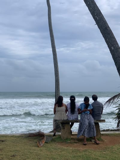 Four individuals, viewed from behind, are seated on a rough-hewn stone bench facing the ocean. Two palm trees frame the scene, one on the left and another on the right, with the sky overhead filled with overcast clouds. The ocean is rough, with white-capped waves crashing against the shore. The foreground consists of a grassy area with some fallen palm fronds. The individuals appear to be a mix of genders and ages, with three women and one man. One woman on the far left is wearing a patterned dress, the woman next to her wears a white patterned top and a light-colored skirt, the woman in the middle is wearing a blue t-shirt and a black and white checkered skirt, and the man on the far right is wearing a patterned shirt and dark pants. The woman in the checkered skirt is looking down at her phone. This scene is located in Koggala, Sri Lanka, a coastal town known for its beaches and natural beauty.
