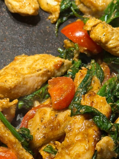 A close-up, overhead shot shows cooked chicken strips, cherry tomato halves, and wilted spinach in a pan. The chicken is golden brown and appears to be seasoned. The tomatoes are bright red and glistening. The spinach is dark green and appears to be cooked down. The pan is dark and textured, suggesting it is a cast iron skillet or similar cooking surface. The lighting is warm and highlights the food. There are no people or any specific location details visible in the image.