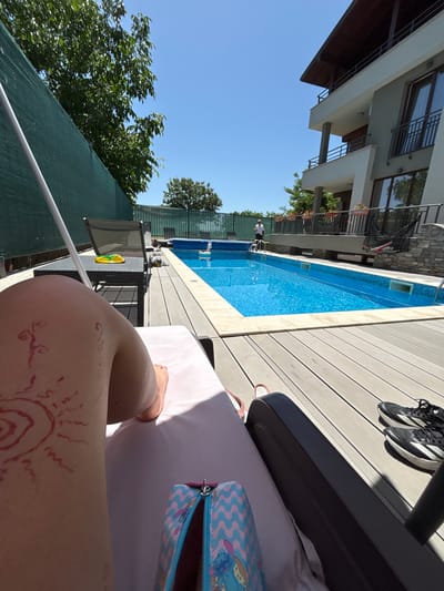 In Ognyanovo, Bulgaria, on a bright sunny day, a person is relaxing on a lounge chair by a swimming pool. The person has red drawings resembling a sun and swirls on their left leg. A colorful pencil case with a Stitch design is on the lounge chair. Black athletic shoes are placed on the wooden deck next to another lounge chair. The swimming pool is blue with a tiled edge and is partially covered. A modern multi-story building with balconies and glass doors is visible in the background, surrounded by trees. A green fence lines the property.