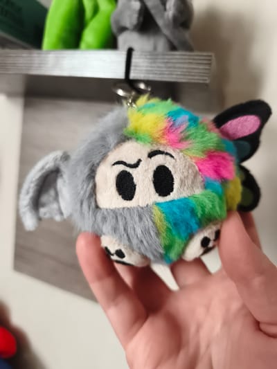 A hand holds a small, fuzzy toy with a gray, elephant-like side and a rainbow-colored, butterfly-winged side. The toy has black button eyes and small black feet. In the background, a shelf holds other out-of-focus toys, one appearing bright green. The scene is indoors, with a plain, light-colored wall visible. The image does not provide any information about the city of Sokolivske, Ukraine.