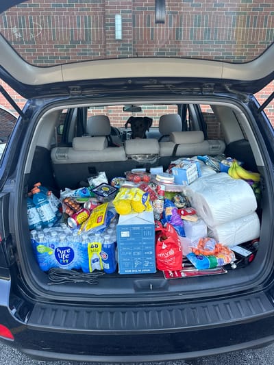 A black SUV is parked in front of a brick building in Prospect Heights, United States of America. The rear liftgate of the SUV is open, revealing the cargo area filled with groceries. A black dog is visible in the front passenger seat of the SUV, looking towards the back. The groceries include numerous cases of bottled water, sports drinks, canned goods, produce such as bananas and carrots, and large packages that appear to be paper towels or similar household items. There are also several bags and boxes of food products. The scene appears to be an outdoor setting, possibly a driveway or parking lot, with a brick wall as the backdrop.