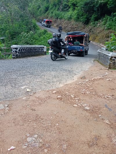 The image shows a paved road in a rural area of Cisewu, Indonesia, with a small bridge in the foreground. Two motorcyclists are riding towards the camera, one behind the other.  The first motorcyclist is wearing a black helmet and dark clothing. The second motorcyclist is wearing a white helmet and a black jacket.  The road is bordered by dense vegetation, and the road surface appears uneven and dusty. In the distance, a blue truck is parked on the right side of the road. There are several people in the truck's bed. The truck has a red canopy and the license plate is visible. There are several cars and motorbikes parked on the left side of the road in the background. The bridge has stone retaining walls and a metal railing.  The scene is likely near the coordinates -7.411, 107.542.  There are no other people present in the image. The sun is shining and the weather is clear.