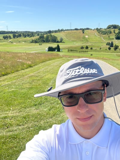 A person wearing a white polo shirt, a gray hat, and black sunglasses takes a selfie on a golf course. The person is positioned in the foreground, with the golf course stretching out behind them. The golf course features rolling green fairways, scattered trees, and a clear blue sky. In the background, power lines and towers are visible. The time of day appears to be daytime, likely sunny, given the clear sky and the shadows. The overall mood seems relaxed and sporty, consistent with a day of golf. The hat has the word "Titleist" embroidered on it in white lettering.