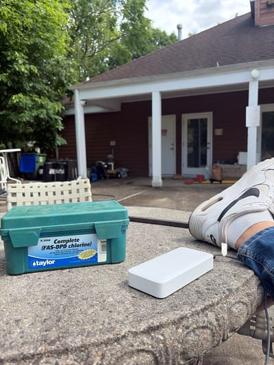The image shows an outdoor setting, likely a backyard or patio area. A leg with a white sneaker and blue jeans is visible in the upper right corner. A white rectangular object, possibly a power bank or similar electronic device, is positioned next to the sneaker. Below the white object is a green container labeled "Taylor Complete (FAS-DPD chlorine) K-2006". The label indicates it's a pool water testing kit. In the background, a house with a brown exterior and white trim is visible. A white plastic chair is partially visible near the testing kit. Green trees are seen on the left side of the frame. The photo could possibly be located in Edison, New Jersey.