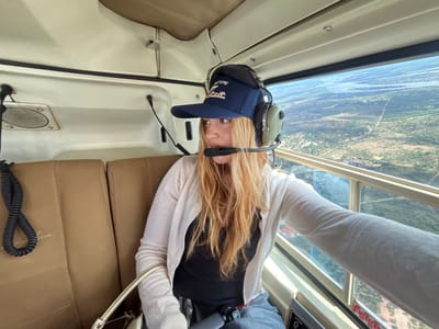 The image shows a woman inside a small aircraft, likely a light plane, flying over Victoria Falls Municipality, Zimbabwe. The woman is wearing a dark blue baseball cap with what appears to be animal silhouettes embroidered on the front, and a headset with a microphone. She is also wearing a white cardigan over a black top. She appears to be a passenger, possibly a tourist, taking in the aerial view.
Outside the plane's window, the landscape of Victoria Falls Municipality is visible, featuring sparse green vegetation and a winding river with braided channels, suggesting a dry season. Scattered buildings, possibly lodges or homes, can also be seen. The sky is hazy. The plane's interior is beige, with a seat next to the woman. An "EMERGENCY" label can be seen on the window frame. The image suggests a tourist activity like a scenic flight over the falls or the surrounding area.