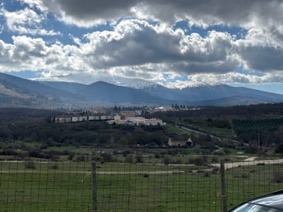 This image presents a panoramic landscape view of the Real Sitio de San Ildefonso, Spain, under a sky exhibiting fragmented cloud cover and visible blue patches. The scene is viewed from behind a wire mesh fence with wooden posts, situated in a green, grassy foreground.
In the background, a substantial mountain range, identified as the Sierra de Guadarrama, spans the horizon, displaying extensive snow-capped peaks and dark, forested lower slopes. Diffused sunlight breaks through the clouds, illuminating parts of the mountains and valley.
The midground features the town of Real Sitio de San Ildefonso, characterized by a cluster of multi-story buildings, predominantly light-colored with red or brown roofs, indicating residential or urban development. To the right of the town, a prominent, light-yellow colored building with a red-tiled roof is discernible. A winding road or highway traverses the valley, running horizontally across the middle of the frame. The landscape between the foreground and the town consists of dark, unfoliated trees or dense shrubbery, alongside terraced agricultural fields, possibly orchards or vineyards, visible on the right side.
No human subjects or specific actions are identifiable within the frame. A portion of a black vehicle, likely a car, is visible in the lower right corner, indicating the immediate vantage point of the photograph.