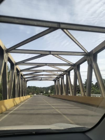 A front-facing, medium shot shows a view from inside a vehicle looking out at a road crossing a bridge. The bridge is constructed of grey steel trusses forming a geometric pattern against a slightly cloudy sky. A yellow concrete barrier lines the right side of the road, which features a white lane divider. The road is paved and appears to be a two-lane road. In the distance, a white vehicle is driving away from the camera. The surrounding environment consists of lush green trees and vegetation on either side of the road, suggesting a tropical or sub-tropical location. The image captures a moment of travel on a sunny day, with the shadow of the bridge's structure cast onto the roadway. The location is identified as Babah Dua, Indonesia.