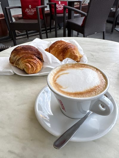 A cup of cappuccino with a white foam surface sits on a matching saucer with a silver spoon resting across it. Beside the cup, two croissants are nestled in a white paper liner. One croissant is golden brown, while the other is darker and appears to have a chocolate filling. The scene is captured on a table, with glimpses of outdoor seating chairs visible in the background.