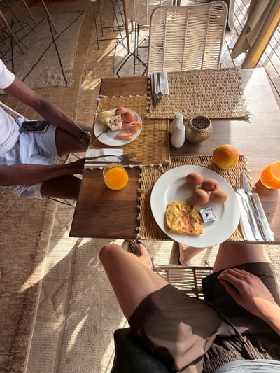 An overhead shot captures a breakfast scene at a resort in Agafay, Morocco, with two individuals present. The setting appears to be an outdoor dining area, suggested by the woven furniture, straw placemats, and the natural light filtering in, casting distinct shadows.

One person, on the left, is seen from the waist down, wearing a white t-shirt and light blue shorts. Their arm is resting on their lap, and a smartphone is placed on top of it.

The other person, on the right, is also viewed from the waist down, wearing brown shorts. Their legs are crossed, and their right hand is resting on their thigh.

On the wooden table, a meal is laid out. A woven placemat holds a white plate with four hard-boiled eggs, a piece of square flatbread, and a small foil-wrapped butter portion. Adjacent to this plate is a whole orange. To the right of the plate is a napkin with a fork and knife.

Another woven placemat on the table displays a glass of orange juice and a small wooden tray with a spoon, a small cup of yogurt topped with granola and fruit, and slices of ham. Further to the right are salt and pepper shakers and a decorative metal object. The background reveals more woven chairs and the textured flooring. The overall impression is a relaxed, sunlit breakfast setting at a Moroccan desert resort.
