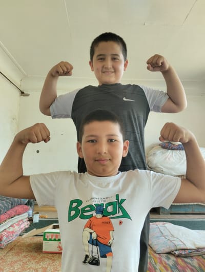Two boys, one in the foreground and one behind him, are posing with their arms flexed. The boy in the foreground is wearing a white t-shirt with a graphic print and the boy in the background is wearing a dark t-shirt with a white stripe and a Nike logo. The boys appear to be in a room with a light-colored ceiling and walls. There are stacks of fabric and some boxes visible in the background, suggesting the location may be a home or a shop. The overall scene is casual and lighthearted.