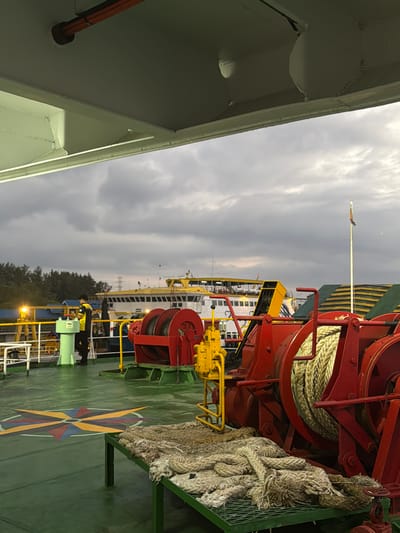 An overcast sky looms over the deck of a ferry in Melaya, Indonesia. In the foreground, thick ropes are coiled on a green metal rack. Behind the ropes, a large red winch with a coiled rope is visible. Further back, a person in a yellow and black uniform stands near a green pillar. Another ferry, with a yellow superstructure and white cabins, is docked in the background. Trees and what appear to be power lines are visible on the shore. The deck has a green painted surface with a colorful compass rose design.