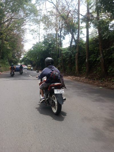 A motorcyclist is traveling on a paved road lined with trees. The trees create a canopy over the road. The rider is wearing a helmet, a blue long-sleeved shirt, and shorts. A black backpack hangs over the rider's shoulder. The motorcycle has a license plate that reads "G 5615 LU 04.29." Other vehicles, including motorcycles and cars, are visible ahead in the distance. The location is identified as Ban Phouviengnoy, Laos.