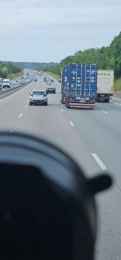 The image depicts a highway scene with multiple vehicles on a cloudy day. In the foreground, the blurry silhouette of a vehicle's interior is visible, suggesting the photo was taken from a car. On the highway, a silver hatchback car is in the left lane, and a blue container truck with "WHEELTAINER" branding is in the right lane, followed by a white truck. Various other cars and trucks are visible further down the highway, indicating moderate traffic. The highway is bordered by green trees on both sides. The context provided suggests this scene is located on a highway near Saint-Cyr-l'École, France, as indicated by the traffic flow and the surrounding vegetation characteristic of French highways. There are no discernible people visible in the image, and the focus is on the vehicular traffic and the highway environment.
