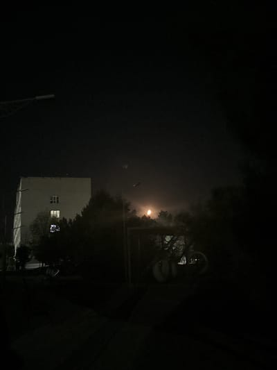This is a nighttime photograph, likely taken outdoors in Egorlykskaya, Russia, given the location context. The scene is predominantly dark, with a sense of mystery and quiet.
The primary subjects visible are a building with lit windows and a streetlamp. The building appears to be a light-colored, multi-story structure. Two windows on its side are illuminated, suggesting occupancy. One of these windows displays a complex, reflective pattern, possibly indicating a decorated interior or a screen reflecting the light. Trees obscure parts of the scene, adding to the nocturnal atmosphere. A tall, slender pole, possibly a utility pole or a light pole, is visible in the lower left quadrant of the image.
The activity taking place is implied rather than explicit. The lit windows suggest normal life occurring within the building, while the streetlamp indicates a public or semi-public space. The overall mood is one of stillness and perhaps isolation, common in nighttime urban or semi-urban environments.
Notable details include the time of day, which is clearly night, as indicated by the darkness and artificial lighting. The presence of fog or mist is suggested by the hazy glow around the streetlamp and the diffuse light in the upper left of the image. This could imply damp weather conditions. The location cues, like the building style and the utility pole, are consistent with a Russian setting.
There is no visible text in the image. The photograph captures a quiet, perhaps slightly eerie, moment in a dimly lit environment.