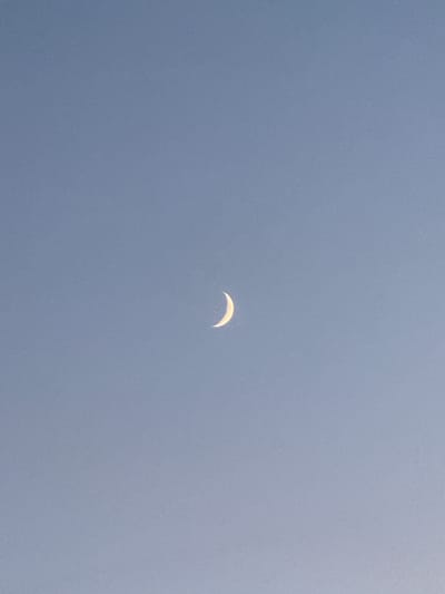 This media file features a crescent moon in a pale blue, overcast sky. The moon is positioned slightly off-center in the frame, appearing as a thin, glowing arc. There are no people, objects, or discernible location cues within the image, creating a minimalist and atmospheric depiction of the celestial body. The lighting suggests it is either dawn or dusk, given the presence of the moon and the muted sky. The overall impression is one of tranquility and vastness, with the sky's texture hinting at a slightly hazy or cloudy atmosphere.