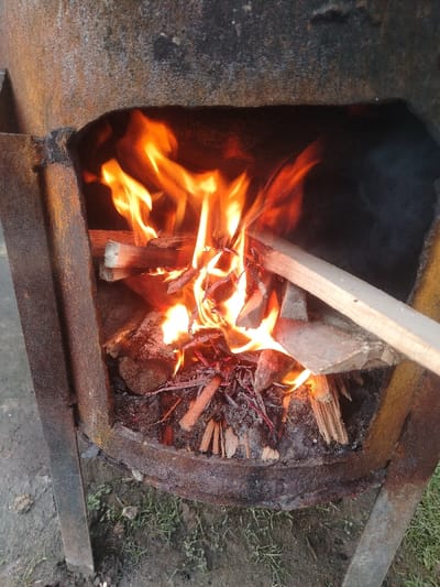 A rusted metal outdoor stove or burner is actively alight, containing a fire fueled by various pieces of wood. Bright orange and yellow flames rise from a bed of burning logs, kindling, and embers, with ash visible at the bottom of the combustion chamber. A flat, light-colored wooden plank extends from the right, partially across the burning material, its end catching fire. The structure itself is a dark, heavy-gauge metal, showing significant rust and wear, and is supported by visible metal legs, one on the left and another partially seen on the right. The stove rests on uneven ground composed of dark soil with scattered patches of green grass or weeds. The immediate area around the fire is illuminated by the flames, while the background is predominantly dark. The scene is located in Nurli zamin, Uzbekistan.
