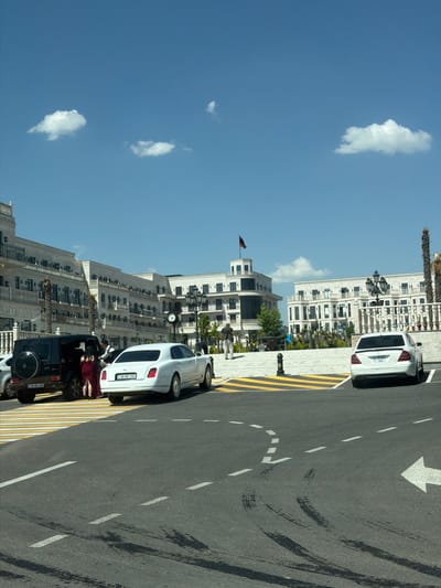 In Proshyan, Armenia, on a sunny day, a dark-colored SUV and a white luxury sedan are parked on a street. The SUV has its rear spare tire visible. A person in a red dress is standing near the SUV, and another person in a dark shirt is by the driver's side of the SUV. The white sedan is positioned behind the SUV. In the background, several multi-story buildings with balconies and decorative facades line the street. Some palm trees are visible in front of the buildings. The sky is blue with a few scattered white clouds. The road has visible tire marks and lane markings, including a white arrow indicating a turn.