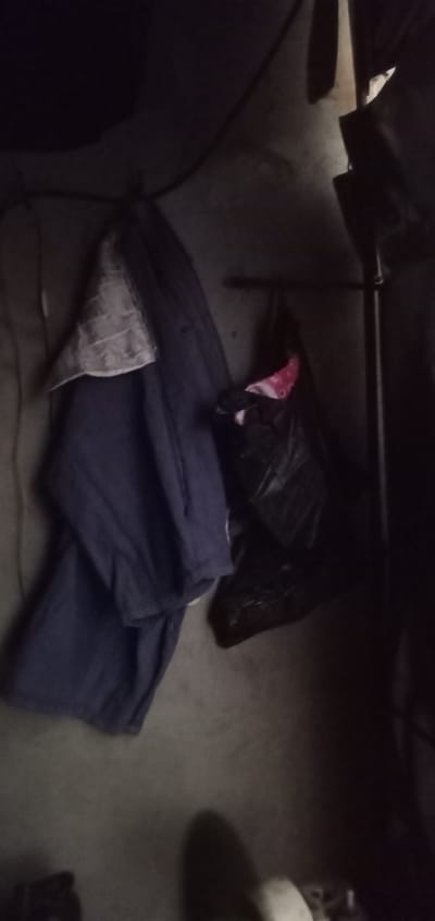The image depicts a dimly lit indoor scene, possibly a room or storage area. On the left, a folded dark blue garment, likely pants or a skirt, is draped over a surface. A lighter colored, possibly patterned fabric is visible within the fold of this garment. To the right of the blue garment, a black plastic bag is hanging, with a portion of pink and white fabric peeking out from its opening. The background appears to be a textured wall or floor, predominantly dark and indistinct. The lighting is low, casting shadows and obscuring fine details. No people are visible in the image. The location is specified as Sumput, Indonesia, suggesting this is an indoor scene within that city.