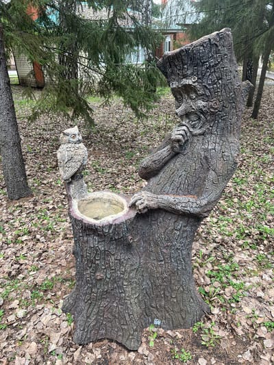 A large, intricately carved wooden sculpture, resembling an old man or forest spirit, stands prominently in an outdoor setting. The sculpture features highly textured surfaces mimicking tree bark, depicting a figure with a long, flowing beard, a hat, and hands placed near a hollowed-out section in its form, which contains some water, possibly serving as a bird bath or planter. An owl sculpture, matching the textured style, is perched on a lower branch-like extension of the main figure.
The ground surrounding the sculptures is covered with a dense layer of dry, fallen leaves, punctuated by small patches of green vegetation. In the background, several coniferous trees, possibly fir or spruce, are visible, creating a wooded atmosphere. Beyond the trees, a light-colored building with a red roof can be partially seen, suggesting the proximity of a residential or institutional structure, consistent with the Sanatoriya Yumatovo Imeni 15-Letiya Bassr location.
The scene is static, with no discernible activity occurring. The lighting is soft and diffuse, indicative of an overcast day or early morning/late afternoon, without strong shadows. The sculptures evoke a sense of folklore and connection to nature. No visible text is present in the image.