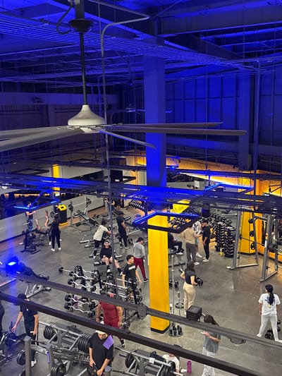 An overhead shot captures a bustling gym atmosphere. Numerous individuals are engaged in weightlifting exercises, utilizing various machines and free weights. The gym is well-lit, featuring a structural ceiling design with visible beams and a large industrial fan. Blue ambient lighting accents the architectural elements, creating a distinct visual ambiance. The overall impression is one of a well-equipped and active fitness center.