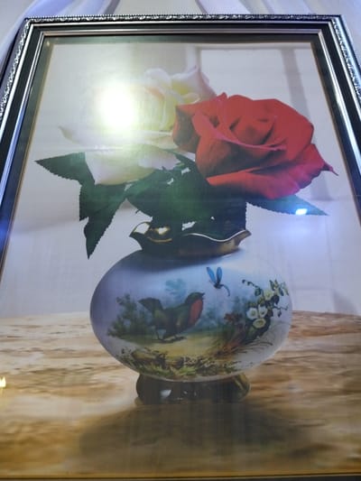 This is a close-up, eye-level, indoor shot of a framed painting. The painting depicts a still life with two roses in a vase. One rose is red, and the other is white or light pink. The vase is white and has a detailed painting of a bird and a dragonfly on it, along with some floral elements. The painting is placed on a reflective surface that appears to be a table with a wood grain pattern. The background of the painting is a muted, light color. The frame of the painting is ornate and dark, with decorative elements along its edges. There is a strong light source reflecting brightly on the painting, suggesting it might be lit by a flash or a nearby lamp. The overall impression is of a decorative artwork. There are no people or specific locations in Cottrueng, Indonesia visible in the image.