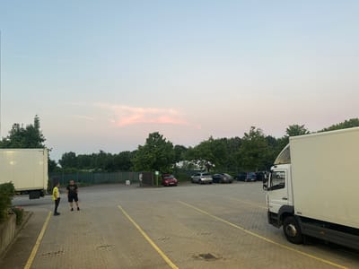 A paved parking lot with yellow lines, located in Schweitenkirchen, Germany, shows two men standing near a white truck. One man, wearing a yellow vest and dark pants, faces the other. The second man, dressed in a black t-shirt and black shorts, faces the man in the vest. Other vehicles, including a red car and several other cars parked in the distance, are visible. A large white truck is parked on the right side of the image. The background consists of trees and a fence. The sky above is a pale blue with hints of pink clouds. A contrail from an airplane is visible in the upper left portion of the sky.