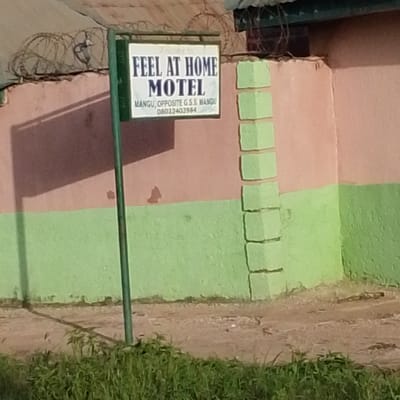 This is an outdoor, eye-level medium shot of a sign for the "FEEL AT HOME MOTEL" in Mangu, Nigeria. The sign is white with dark blue text and is mounted on a green pole. The text on the sign reads: "Welcome to FEEL AT HOME MOTEL MANGU, OPPOSITE G.S.S. MANGU 08032403964". The motel building behind the sign is painted in two horizontal bands, the upper band pink and the lower band green. To the right of the sign, the pink wall is decorated with a vertical series of green rectangular blocks. Above the pink wall, barbed wire is visible. The ground in front of the building is dirt with some patches of green grass in the foreground. The lighting suggests it is daytime, with shadows cast on the wall. The scene is a typical roadside commercial establishment in Nigeria.