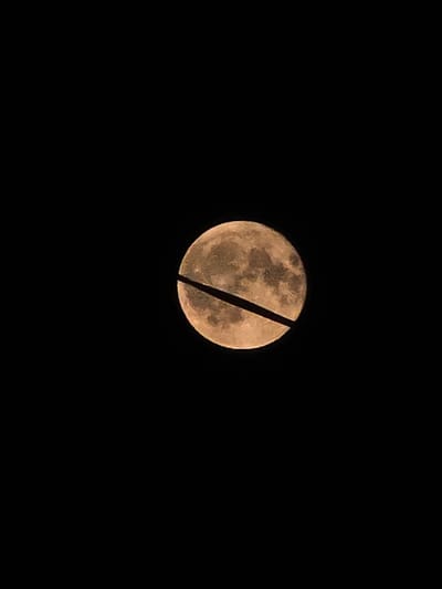 A full moon, partially obscured by a black line, is visible in a dark sky. The moon has a yellowish-orange hue and displays visible surface textures. The black line appears to be a silhouette of a power line or similar structure passing horizontally across the face of the moon. The overall composition is minimalistic, focusing entirely on the celestial body and the obstructing element. There are no visible signs of the city of Khojniki, Belarus, in this image.