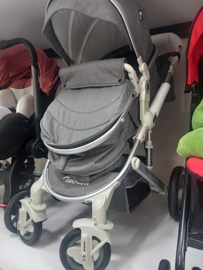 A close-up, low-angle shot shows a grey stroller with a white frame in a retail environment, possibly a store in Khodja Akhrar Vali, Uzbekistan. The stroller is the primary subject, positioned centrally. It features a quilted grey fabric seat and canopy, with a silver trim around the seat. The frame is white, with exposed suspension springs visible on the rear supports. Part of a red stroller is visible to the right, and another stroller with a red lining is visible to the left, suggesting a display of multiple baby products. The floor appears to be white. The lighting is bright, casting shadows on the floor. The scene is static, with no people or ongoing actions visible.