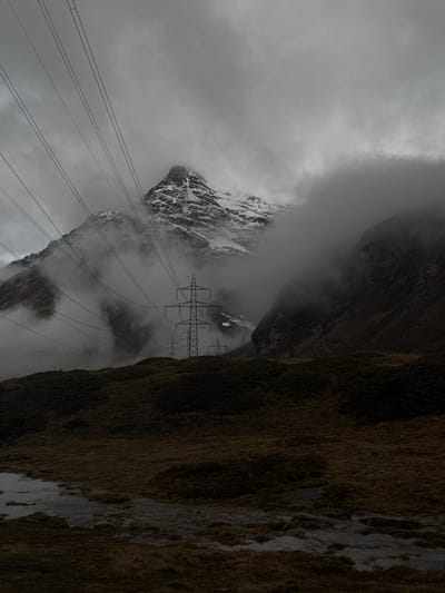 The image presents a dramatic and rugged mountainous landscape, likely situated in the Swiss Alps near Pontresina, captured under heavily overcast and foggy conditions. The scene is dominated by a steep, dark, and rocky mountainside on the right, covered in sparse, dark vegetation and featuring vertical streaks of water or melted snow.
In the mid-ground and background, snow-capped peaks emerge from dense, swirling grey clouds and fog, which obscure their upper sections and create a sense of depth and mystery. Cutting across the frame, multiple parallel power lines stretch across the mountainous terrain. Several large electricity transmission towers (pylons) are visible, with some partially shrouded by the low-hanging clouds.
The weather appears cold and damp, with a muted light characteristic of an overcast day. The predominant colors are somber greys of the clouds and snow, dark browns and greens of the rocky slopes, and the stark black of the power lines and towers. No human activity or individuals are visible, nor is any text present in the image. The scene portrays a blend of natural alpine grandeur and human infrastructure.