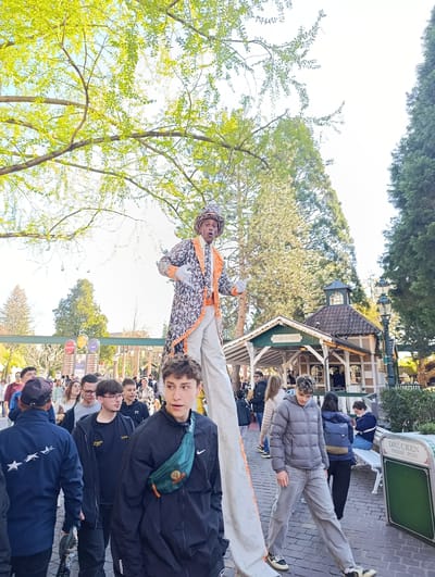 The image depicts a street scene in Rust, Germany, likely within or near the Europa-Park amusement park, due to the presence of the "Panorama-Bahn" station in the background. A person on stilts, dressed in a patterned coat, white pants, and a hat, is the central focus, performing or interacting with the crowd. The crowd consists of a diverse group of people, including teenagers and adults, some of whom are looking at the performer. The foreground features individuals walking towards the viewer. The environment includes trees, buildings resembling park structures, streetlights, benches, and signage (one indicating "Drücken Presse Push"). The lighting is bright, suggesting daylight, with tree branches and leaves visible overhead.