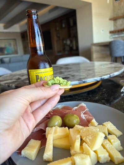 A hand holds a cracker topped with green dip next to a bottle of Pacifico beer. A gray plate on a counter features thin slices of prosciutto, green olives, and chunks of Parmesan cheese. The counter is black with a patterned edge. A glimpse of the interior of a house is visible in the background.