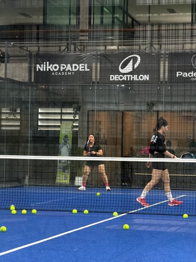 This is a medium shot photograph taken indoors, likely in a Padel academy or club, located in Ban Buak Khrok North, Thailand. The scene depicts a Padel court with a blue surface and white lines. Several yellow Padel balls are scattered on the court. A white net divides the court.
In the foreground, on the left side, is the lower body of a person wearing black shorts, pink socks, and white sneakers. On the right side, a person is walking across the court, wearing beige shorts, white socks, and red sneakers. They are also wearing a black t-shirt with white writing on the back.
In the mid-ground, another person is standing on the left side of the court, wearing black shorts and a black t-shirt, holding a Padel racket. Behind them, a green banner with text and an image is visible.
In the background, through the glass walls of the court, there are signs visible. One sign reads "Niko Padel Academy". Another sign has the "DECATHLON" logo and text. Further to the right, a sign reads "Padel". Reflections of lights and architectural elements are visible on the glass. The overall lighting appears to be artificial, common for indoor sports facilities.