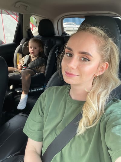 A woman takes a selfie inside a car, with a young child visible in a car seat behind her. The image is captured from an upward angle, showing the woman's face and upper torso in the foreground, turned slightly towards the camera. She has long blonde hair, styled partly in a ponytail, and is wearing a plain green t-shirt and a dark seatbelt across her chest. She has makeup on, including winged eyeliner, and a neutral expression with a slight smile. A small golden hoop earring is visible on her left ear.
Behind her, to the right, a toddler is seated in a dark-colored car seat. The child is wearing a leopard-print top, ribbed brown leggings, and white patterned socks. They are looking forward, with a neutral expression. A yellow warning label with the word "AIRBAG" is visible on the side of the car seat.
The car's interior features dark upholstery. Through the car windows, exterior elements are faintly visible, including what appears to be a structure with red and white components, suggesting a daytime setting. The scene takes place in Kiparisovo-2, Russia.